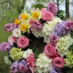 A photo of a wreath made of vibrant spring flowers. The wreath is placed on a wooden door. The flowers include bright yellow daffodils, pink and white roses, purple lilacs, and white and green hydrangeas. The background is a lush green forest. The lighting is soft.
