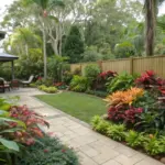 A shaded Queensland garden sanctuary featuring tropical shade plants arranged along a winding stone pathway. Native ferns, palms, and diverse foliage thrive beneath a wooden pavilion, creating a peaceful retreat typical of subtropical shade gardens in Queensland. The well-maintained path leads through layers of lush greenery, demonstrating ideal plant placement for shaded garden spaces.