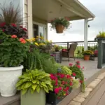 Container plants for front porch create a welcoming display, featuring a mix of potted flowers and greenery in varying sizes. Pink, yellow, and red blooms complement lush foliage arranged along a wooden deck with railing. The well-curated collection of container plants frames a scenic view of open fields under cloudy skies, adding natural charm to the covered porch setting.