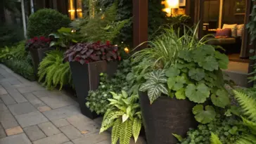 Container plants for shade thrive in this lush garden setting, featuring potted ferns and various greenery arranged along a stone-paved walkway. The garden borders a building whose warm interior lights cast a golden glow through windows, illuminating the shade-loving container garden in the evening hours.