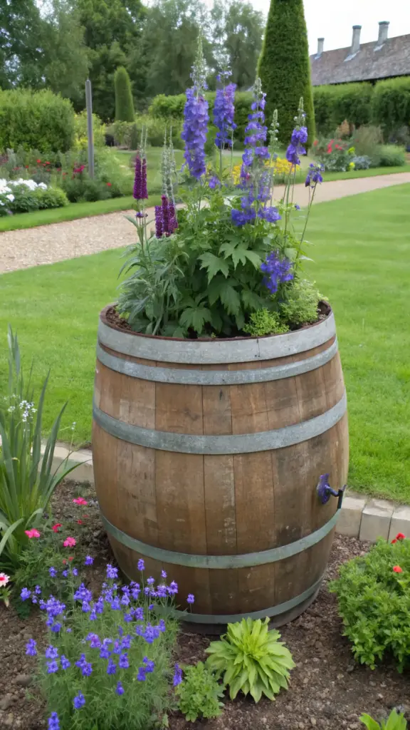 A wooden rain barrel serves as a vibrant cut flower garden container, filled with flourishing purple and blue delphiniums amidst greenery. Positioned on dark brown soil, the light brown barrel features metal bands around its midsection. Surrounding flowerbeds showcase a mix of purple, blue, and pink blooms, complemented by small gravel pathways. Beyond the garden, manicured lawns and pruned trees create a well-maintained backdrop under sunny skies.