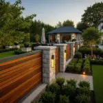 A beautifully landscaped backyard at dusk, featuring a modern garden fence with horizontal wooden slats and elegant stone pillars. The fence posts are adorned with illuminated sconces, and the pathway is lit with ground-level lights, showcasing appealing "Garden Fence Ideas."