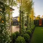 Lush, modern backyard with a wooden deck and a privacy fence. The fence is composed of tall bamboo stalks covered in climbing white flowers, small manicured shrubs, and a slatted black fence with a decorative rusted metal panel featuring a cutout geometric pattern. The keyword "Privacy Fence Ideas" is highlighted in the description.