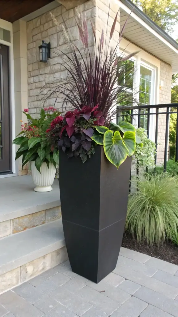 A tall, modern black planter brimming with a dramatic arrangement of dark purple fountain grass, coleus, and vibrant green elephant ear, serving as a stylish example of full sun planter ideas for a home's entryway, with a smaller white planter and stone facade in the background.