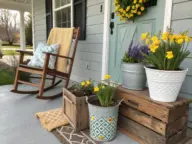 A cozy front porch decorated with Spring Porch Ideas, featuring a wooden rocking chair with a yellow throw and floral pillow, a teal door with a yellow wreath, and various potted daffodils and lavender arranged on rustic wooden crates.
