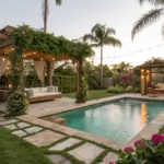 A luxurious swimming pool surrounded by Dreamy Backyard Ideas, featuring a greenery-covered pergola with a white daybed, warm string lighting, and tropical palm trees at sunset.